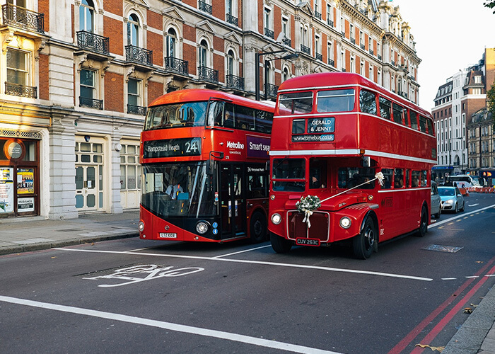 Red double-decker buses in London, featuring a modern bus with route number 24 and a vintage wedding bus adorned with flowers, set against historic architecture.