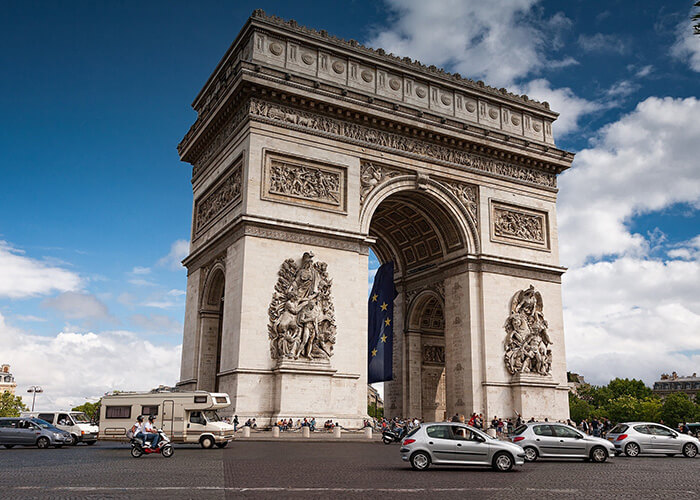 Arc de Triomphe in Paris, showcasing intricate sculptures and European flag, surrounded by bustling traffic and pedestrians.